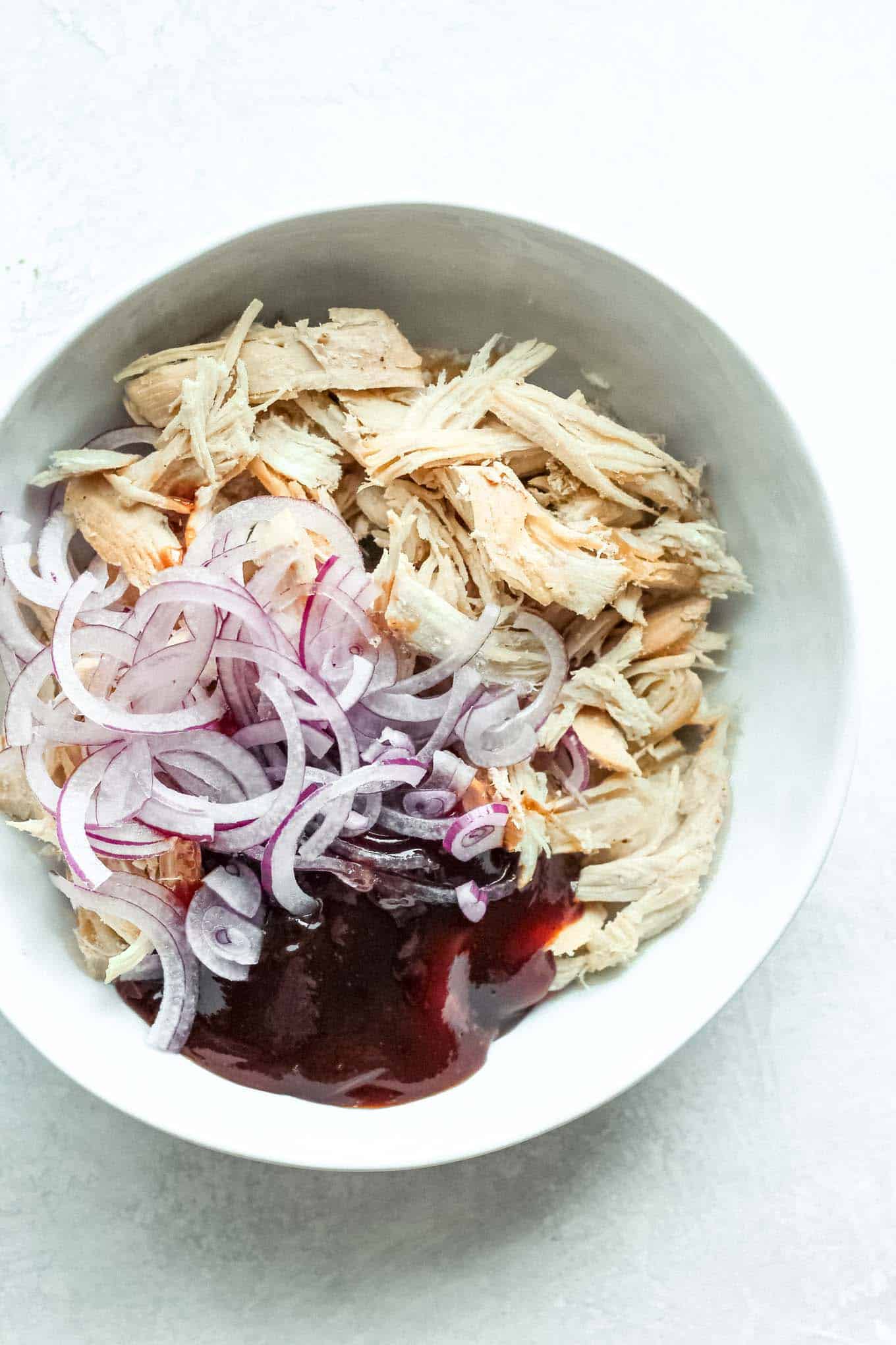 A bowl containing shredded chicken, BBQ sauce, and red onion before mixing