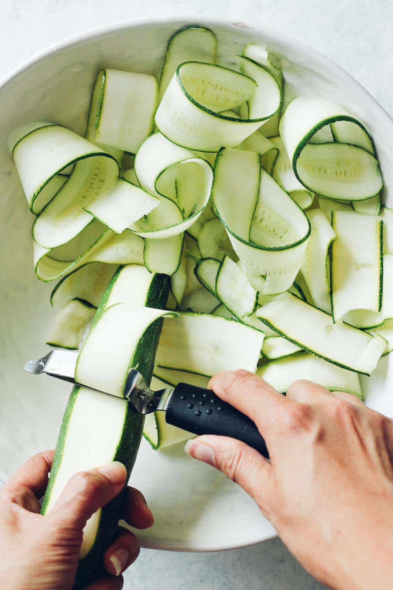 Using a vegetable peeler to shave zucchini ribbons into a large salad bowl
