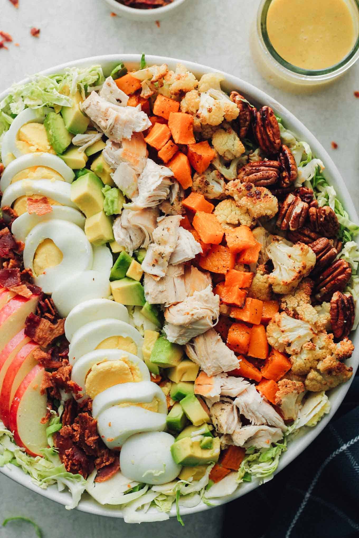 A zoomed in view of the finished Cobb salad in a bowl with the dressing, extra bacon bits, and pecans on the side.