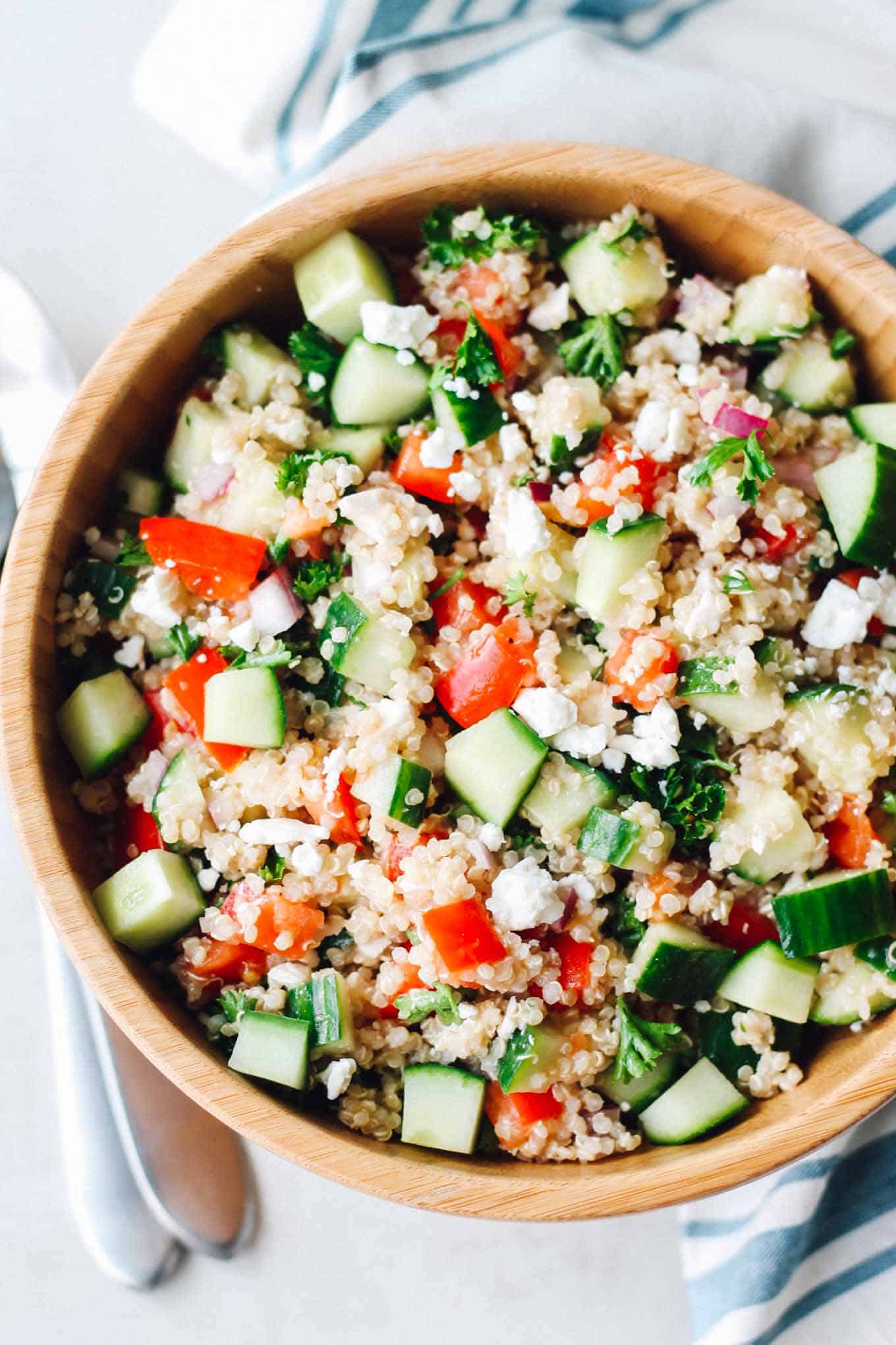 Greek Quinoa Salad in a wooden bowl with a napkin and serving utensils on the side.